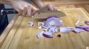 Person slicing red onion on a wooden cutting board with a knife.